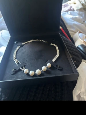 Maria Nilsdotter halsband  - Unikt halsband med grov silverfärgad kedja, dekorerat med fem vita pärlor och två säkerhetsnålar som edgy detalj. Perfekt statement-piece för dig som gillar att sticka ut och mixa klassiskt med punk. Snyggt att bära till både t-shirt och skjorta.