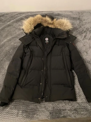 Svart dunjacka från Canada Goose - Svart dunjacka från Canada Goose med fluffig päls på huvan och klassisk logga på ärmen. Jackan har dragkedja, knappar och två stora fickor framtill. Perfekt för kalla vinterdagar med sin tjocka, quiltade design och snygg passform.  Jackan är självklart äkta