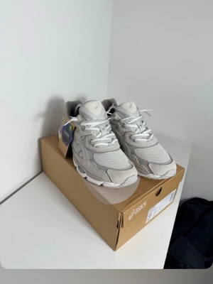 Vita och grå Asics sneakers - Fräscha Asics sneakers i vit och ljusgrå färg med mesh och mockadetaljer. Skorna har snörning, Ortholite-innersula och en chunky platt sula. Perfekt för dig som gillar sportig stil och vill ha bekväma sneakers med snygg design.