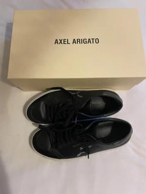 Svarta sneakers Axel Arigato - Snygga svarta sneakers från Axel Arigato med vit sula och svart snörning. Skorna har en stilren design med fågelbroderi på sidan och textdetaljer. Tillverkade i skinn för en exklusiv känsla. Perfekta för dig som gillar minimalistisk och modern stil.