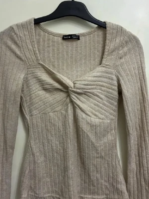 Beige ribbad topp med twist från SHEIN - Snygg beige ribbad topp från SHEIN i storlek XS. Toppen har lång ärm, twistad detalj framtill och en fyrkantig ringning. Materialet är mjukt och stretchigt, perfekt för dig som gillar en slim passform. Den neutrala färgen gör den enkel att matcha med allt.
