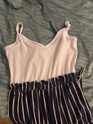 Randig blåvit byxdress med knytband - Snygg byxdress med vit överdel och blå shorts med vita ränder. Byxdressen har smala axelband och ett elastiskt midjeband med knytband i sidan. Materialet känns mjukt och luftigt, perfekt för varma dagar. Ränderna ger en fräsch och trendig look.