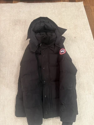 Svart dunjacka från Canada Goose - Säljer en svart dunjacka från Canada Goose med huva och klassisk logga på ärmen. Jackan är tjockt vadderad, har dragkedja och knappar framtill samt två stora fickor. Perfekt för kalla dagar.