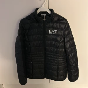 Svart pufferjacka från Emporio Armani EA7 - Svart pufferjacka från Emporio Armani EA7 med logga på bröstet. Jackan har huva, dragkedja framtill och quiltad design. Perfekt för kyliga dagar och har en clean, sportig look. Materialet är syntet och jackan är lätt och smidig att bära. Ordinarie 2000kr pris 1500kr, dragkedjan är lite trasig men går att använda