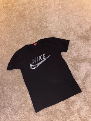 Svart Nike t-shirt med tryck - Svart t-shirt från Nike i storlek L med ett stort, grått Nike-logotryck på bröstet. Trycket har en cool, sprucken effekt som ger en unik vibe. Klassisk rund hals och korta ärmar. Perfekt för dig som gillar sportiga och stilrena plagg.