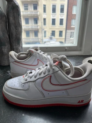 Nike Air Force 1 vit/röd sneakers - Säljer ett par klassiska Nike Air Force 1 sneakers i vitt skinn med röda detaljer och röd yttersula. Skorna har perforerad tå, vit snörning och den ikoniska swoosh-loggan med röd kant. Perfekt för dig som vill ha en clean men ändå unik look.