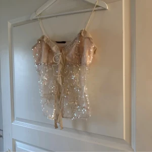 Glittrig beige korsettopp Emmabelin XS-S - Supersöt glittrig korsettopp från Emmabelin i beige mesh med paljetter. Toppen har tunna axelband, volangkant upptill och knytband framtill. Transparent och croppad modell som ger en trendig och feminin vibe. Perfekt för dig som vill sticka ut!