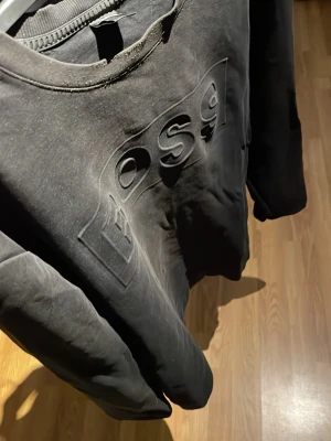 Svart sweatshirt från Boss - Säljer en svart sweatshirt från Boss med stor, upphöjd logga på bröstet. Tröjan har rund halsringning och långa ärmar. Perfekt för dig som gillar stilrena och klassiska plagg med en twist. Passar till det mesta och är riktigt skön.