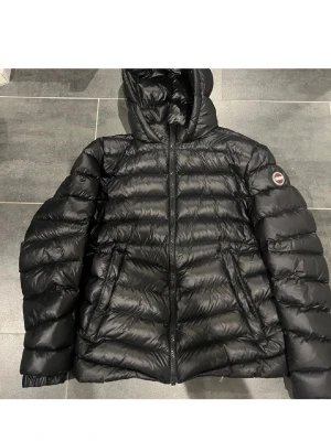 Svart Colmar dunjacka  - Säljer en svart dunjacka från Colmar Originals med huva och dragkedja samt Colmar-logga på ärmen. Perfekt för kalla dagar och har en snygg, puffig look. Jag är 174 och väger 75kg och den passar bra! Men en liten skada på vänstra underarm, detta är dock lagat så synd knappt. (pris kan diskuteras)