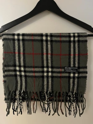 Burberry Halsduk - Snygg rutig halsduk från Burberry i 100% lammull, med klassiskt mönster i grått, svart, vitt och röda linjer. Halsduken har fransar nedtill och en mjuk känsla. Perfekt för kallare dagar! Skriv vid minsta fundering!
