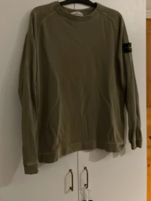 Beige Stone Island sweatshirt - Säljer en beige sweatshirt från Stone Island med klassisk patch på ärmen. Tröjan har rund hals, ribbade muddar och är tillverkad i mjuk bomull. Perfekt för dig som gillar streetwear och vill ha en clean look. Tyvärr lite sliten på ärmen men inget man tänker på.