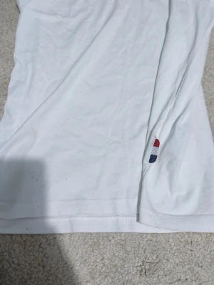 Vit Moncler t-shirt med broderad detalj - Vit t-shirt från Moncler i 100% bomull. Klassisk passform med korta ärmar och rund hals. Snygg broderad detalj i rött, vitt och blått vid nederkanten samt randig insida på kragen. Perfekt för en clean och stilren look.