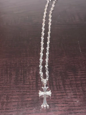 Chrome hearts kedja med korshänge - Säljer en snygg silverfärgad kedja med detaljerade länkar och ett korsformat hänge. Kedjan har små blommor längs hela längden och ett klassiskt T-lås. Korset har en cool, stilren design med rundad mitt och böjda kanter. Perfekt accessoar för att lyfta din outfit.