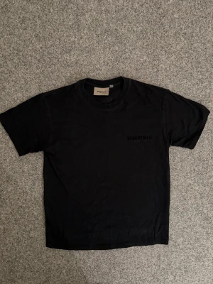 Svart Essentials t-shirt i bomull - Svart t-shirt från Essentials med diskret logga på bröstet och en patch med logga i nacken. Klassisk rund halsringning och korta ärmar. Tillverkad i mjuk bomull för en skön känsla och clean look.