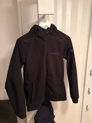 Svart vindjacka från Sail Racing - Svart vindjacka från Sail Racing med huva samt en rund logodetalj på ärmen och huvan. Nypris ca2000kr