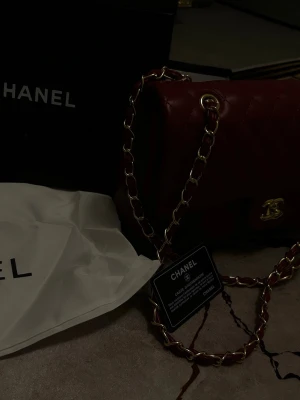 Röd quiltad axelväska  - Snygg röd axelväska  i quiltat skinn med gulddetaljer. Väskan har en klassisk design med en ikonisk logotyp på framsidan och en praktisk dragkedja inuti. Perfekt för att ge en touch av lyx till din outfit.