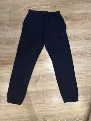 Svarta mjukisbyxor från Replay - Snygga svarta mjukisbyxor från Replay med elastisk midja och snörning. Byxorna har en röd broderad R-logga på sidan och är gjorda i mjukt bomullsmaterial. Passformen är normal med smalare ben och muddar vid bensluten. Perfekta för chill eller träning.