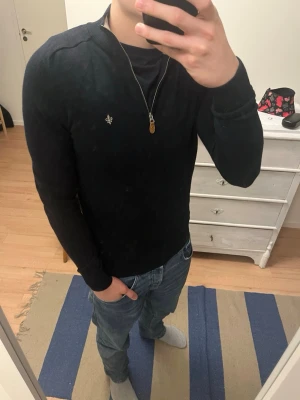 Svart/mörkblå half zip tröja Morris M - Svart långärmad tröja från Morris med beige krage och half zip-dragkedja. Tröjan är stickad i 100% merinoull och har en liten broderad logga på bröstet. Perfekt för dig som gillar stilrena och klassiska plagg med snygga detaljer. 