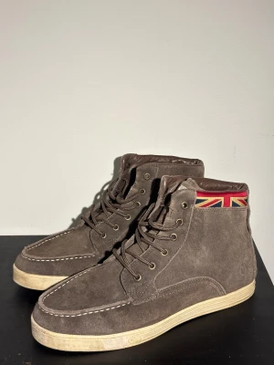 Bruna mocka boots med Union Jack-detalj - Snygga bruna boots i mocka med högt skaft och snörning. Skorna har en beige platt sula och vita sömmar som kontrast. På skaftet finns en cool Union Jack-detalj i rött, blått och gult som ger extra edge. Perfekt för dig som gillar brittisk stil och vill sticka ut.