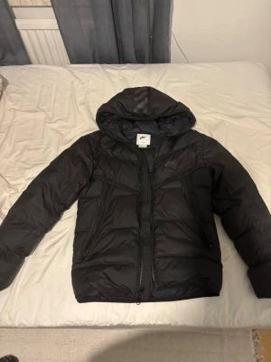 Svart Nike Air Max pufferjacka med huva - Svart pufferjacka från Nike Air Max med stor huva och två snedställda dragkedjefickor framtill. Jackan har AIR MAX-tryck på ryggen och Nike-logga på bröstet. Perfekt för kalla dagar, med stilren look och skön passform.