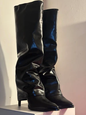 Svarta höga stövlar med klack | boots - Säljer ett par svarta, glansiga stövlar med spetsig tå och låg/medel klack (8cm) Stövlarna har en slouchy, rynkad design och är tillverkade i syntetmaterial som ger en cool och edgy look. Perfekta för dig som vill sticka ut med en statement-sko.