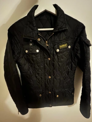 Svart quiltad jacka från Barbour - Quiltad svart jacka från Barbour International med gulddetaljer och dragkedja framtill. Jackan har klassisk krage, tryckknappar och Barbour-logga på bröstet. Snygg passform och långärmad design, perfekt för höst och vår.