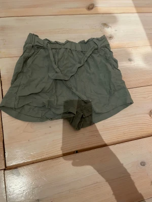 Gröna shorts med knyt från KappAhl - Snygga gröna shorts från KappAhl i storlek 140. De har elastisk midja och ett matchande knytband framtill. Lätta och sköna, perfekta för varma dagar. Stilen är enkel och avslappnad, passar dig som gillar en chill look.