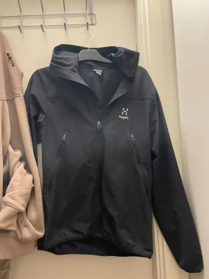  Haglöfs jacka  - Svart vindjacka från Haglöfs med huva och dragkedja framtill. Jackan har två sidofickor med dragkedja och en stilren design med logga på bröstet. Perfekt för blåsiga dagar och outdoor-aktiviteter. Långärmad och tillverkad i slitstark syntet.