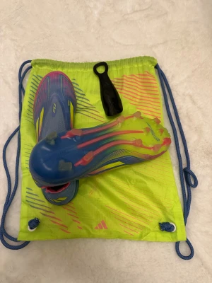 Adidas F50 blå och rosa fotbollsskor - Säljer ett par Adidas F50 fotbollsskor i blått och rosa med gula detaljer. Skorna har en strömlinjeformad design, platt sula med dobbar och är tillverkade i syntetmaterial. Kommer med en neonfärgad skopåse och skohorn. Perfekt för dig som vill sticka ut på planen.