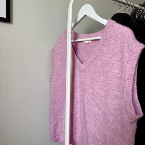 Stickad rosa väst från H&M - Mysig stickad väst i ljusrosa från H&M. Modellen har v-ringning, ribbade kanter och är ärmlös. Perfekt att slänga över en skjorta eller t-shirt för en trendig lager-på-lager-look.