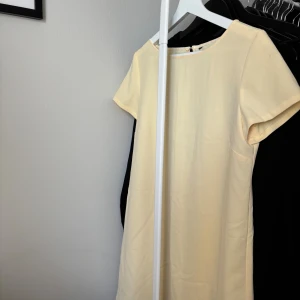 Ljusgul kort klänning från Zara - En stilren ljusgul klänning med kort ärm och rund halsringning. Klänningen är rak i modellen och slutar ovanför knäna. Den har en diskret dragkedja bak och är tillverkad i ett mjukt, slätt tyg som ger en clean look. Från Zara, kortare klänning, slutar ovanför knäna.