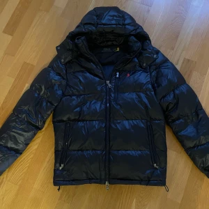 Mörkblå pufferjacka från Polo Ralph Lauren - Säljer en mörkblå pufferjacka från Polo Ralph Lauren med huva och dragkedjor. Jackan är köpt förra vinter och är i väldigt bra skick, finns inga skador på den. Perfekt för kalla dagar och riktigt snygg passform.