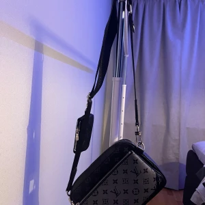 svart axelväska - Snygg svart axelväska  med klassiskt monogrammönster i grått och svart. Väskan har justerbar axelrem med logotyp, silverfärgade dragkedjor och en liten avtagbar plånbok på remmen. Perfekt storlek för mobil och småprylar.