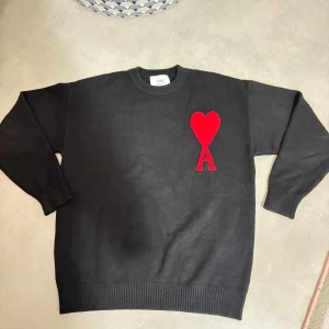 Svart stickad tröja från Ami Paris - Svart stickad tröja från Ami Paris med rund hals och ribbade muddar. På bröstet finns ett stort rött hjärta med bokstaven A, vilket ger en unik och cool detalj. Perfekt för dig som gillar stilrena plagg med en twist.