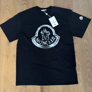 Svart Moncler t-shirt med stor logga - Svart t-shirt från Moncler med stor, cool logga i silver på bröstet och ett broderat Moncler-märke på ärmen. Materialet är mjuk bomull som känns skönt mot huden. Perfekt för dig som gillar streetwear och exklusiva märken.