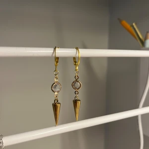 Guldiga örhängen med kristall och kon - Snygga örhängen i guldton med hängande konformad detalj och en klar kristall ovanför. Perfekt för dig som gillar statement-smycken med lite edge. De har en enkel krok för att sätta i örat och passar till många olika stilar. I rostfritt stål och handgjort