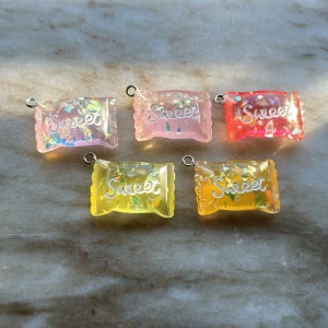 Färgglada godisberlocker med texten Sweet - Fem söta berlocker formade som godis, med texten 'Sweet' på varje. Kommer i färgerna rosa, röd, gul och orange med glittriga detaljer. Perfekta att använda till halsband eller armband för en lekfull och unik stil.
