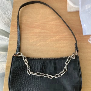 Svart handväska med kedja från pull&bear - Snygg svart handväska från Pull&Bear med croco-mönstrad yta och silverfärgad kedjedetalj. Väskan har dragkedja upptill och ett extra fack med dragkedja på baksidan. Man kan se att den är lite rostigt men det är okej funkar vanligt som en väska ❤️