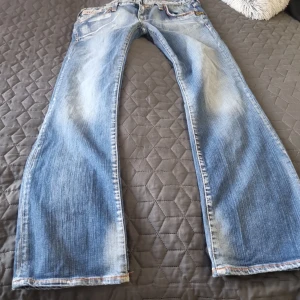 Blå jeans från G-Star med broderi - Snygga blå jeans från G-Star med slitna detaljer och broderad logga på baksidan. Modellen har raka ben, orangea sömmar och klassisk femficksdesign. Jeansen är tillverkade i denim och har en cool, lite vintage tvätt. Är inte säkert på storleken men jag tog midjemått är I storlek 40. 