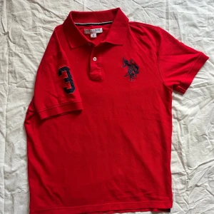 Röd polo t-shirt från U.S. Polo Assn. - Klassisk röd pikétröja från U.S. Polo Assn., herrstorlek S. Tillverkad av mjuk och slitstark bomull med sportiga detaljer: • Stor broderad logotyp på bröstet • Nummer 3 på höger ärm • Klassisk krage med knappslå  📏 Mått (platt): • Längd: ca 62 cm • Bröstvidd: ca 50 cm  Skick: Mycket bra – inga fläckar eller hål. Perfekt för både vardagsbruk och smarta casual-tillfällen.