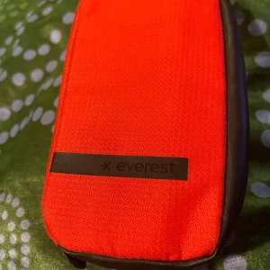 Röd mobilväska från Everest - Snygg och kompakt röd mobilväska från Everest med svart kant och praktiskt innerfack. Väskan är tillverkad i slitstark polyester och har dragkedja upptill. Perfekt för att skydda och bära mobilen till skolan eller jobbet.