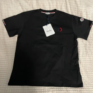 Svart t-shirt från Moncler med logga - Snygg svart t-shirt från Moncler med broderad logga i rött och blått på bröstet samt patch på ärmen. Klassisk passform och rund hals. Små detaljer i rött, vitt och blått på ärmarna. Top kvalité.