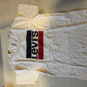 Vit Levi's t-shirt med logga - Vit t-shirt från Levi's i storlek L med klassisk tryckt logga i svart, blått och rött på bröstet. Modellen har rund hals och korta ärmar. Tillverkad i mjuk bomull som är skön mot huden. Perfekt för dig som gillar streetwear och enkel stil.