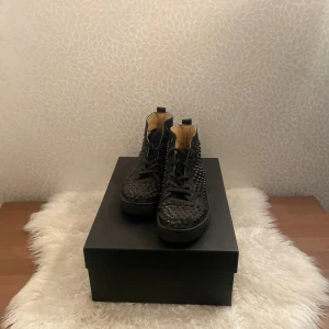 Svarta Louboutin sneakers med nitar - Säljer ett par svarta high-top sneakers från Christian Louboutin med ikonisk röd sula och coola nitar över hela skon. Skorna har snörning och är tillverkade i skinn med en platt sula. Perfekt för dig som vill sticka ut med en edgy vibe. Pris går att diskutera 