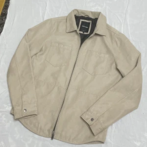 Jacka  - Snygg beige overshirt från Jack & Jones i storlek L. Jackan har två bröstfickor, klassisk krage och dragkedja framtill. Materialet är mockaliknande syntet med svart foder på insidan. Perfekt för lager-på-lager och chill stil.