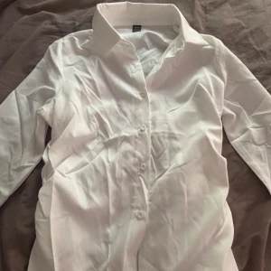 Vit klassisk skjorta med knappar - Snygg vit skjorta med klassisk krage och knappar framtill. Skjortan har lång ärm. Perfekt för dig som gillar stilrena och enkla plagg. Passar dig som vill ha en clean look.