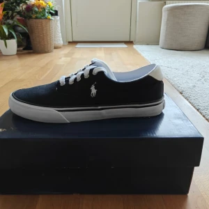 Svarta sneakers från Polo Ralph Lauren - Stilrena svarta sneakers från Polo Ralph Lauren med vit sula och vita detaljer. Skorna har klassisk snörning, rund tå och är tillverkade i textil. Ikoniska Polo-loggan broderad på sidan och Polo-tryck på hälen. Perfekta för en clean och avslappnad look.