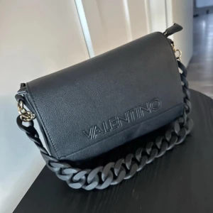 Svart axelväska från Valentino - Med det extra bandet 480kr och bara den svara 380kr. Så skriv om ni är intresserade                                                                      Snygg svart axelväska från Valentino med präglad logga på framsidan. Väskan har en bred kedjerem i plast och en extra justerbar rem med logotyp. Stilren och modern design i skinnimitation, perfekt för att lyfta din outfit.