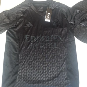 Svart Real Madrid fotbollströja Adidas - Snygg svart Real Madrid fotbollströja från Adidas med diskret mönster och klubbmärke broderat på bröstet. Tröjan har korta ärmar, V-ringad krage och är tillverkad i lätt, ventilerande Aeroready-material. Perfekt för dig som vill ha en stilren look på planen.