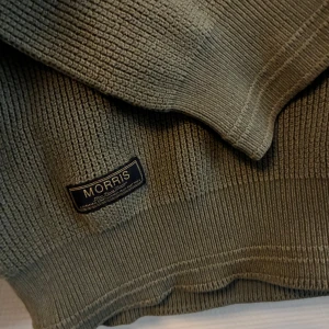 Olivgrön stickad tröja från Morris - Olivgrön stickad tröja från Morris med broderad logga på bröstet. Tröjan har rund halsringning, ribbade muddar och en klassisk passform. Perfekt för lager-på-lager och snygg till jeans eller chinos. Passar nog även xs.